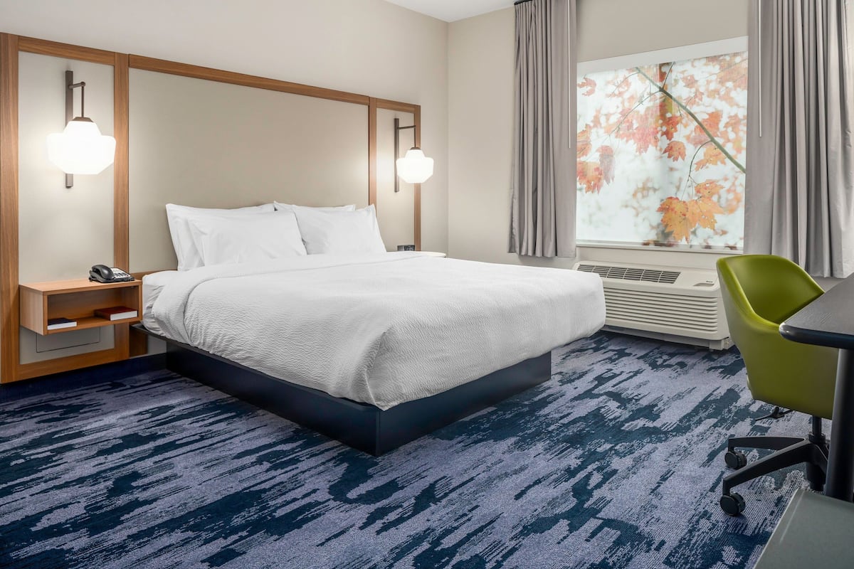 A hotel room with a bed, bedside tables, a desk with a chair, a window with curtains, and a wall-mounted air conditioner.