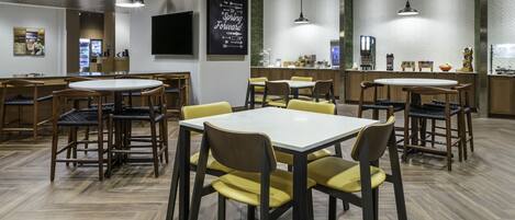 A modern restaurant with round tables and chairs, a kitchen area, and a herringbone-patterned wall.