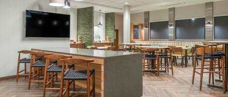 A modern restaurant with a central bar area, multiple seating options, and a large flat-screen TV mounted on the wall.