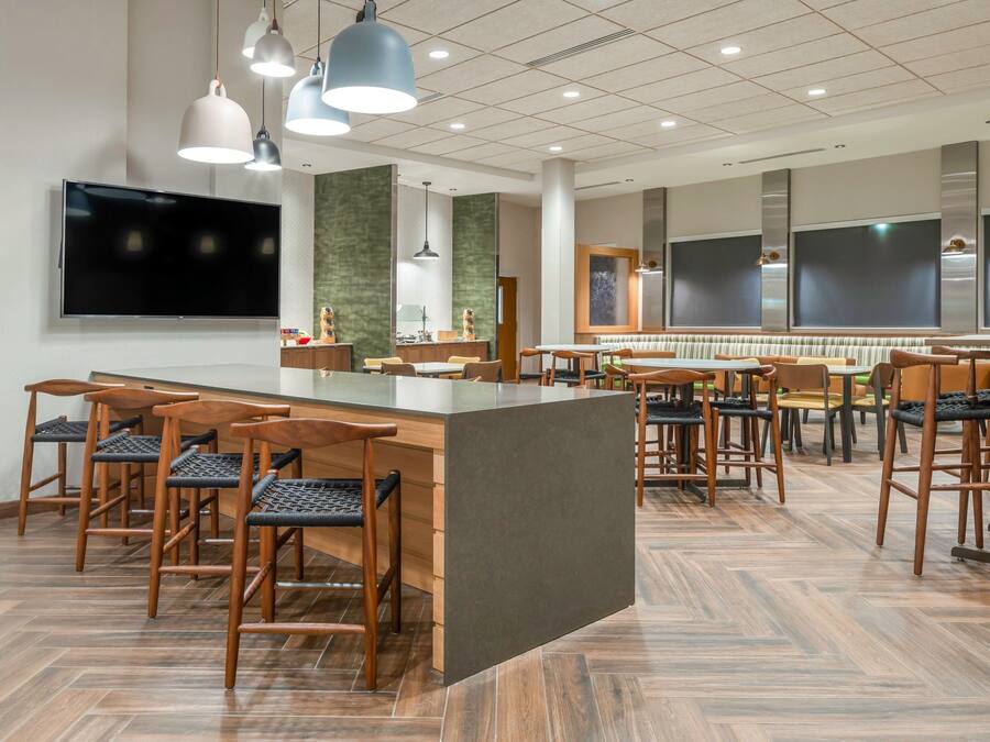 A modern restaurant with a central bar area, multiple seating options, and a large flat-screen TV mounted on the wall.