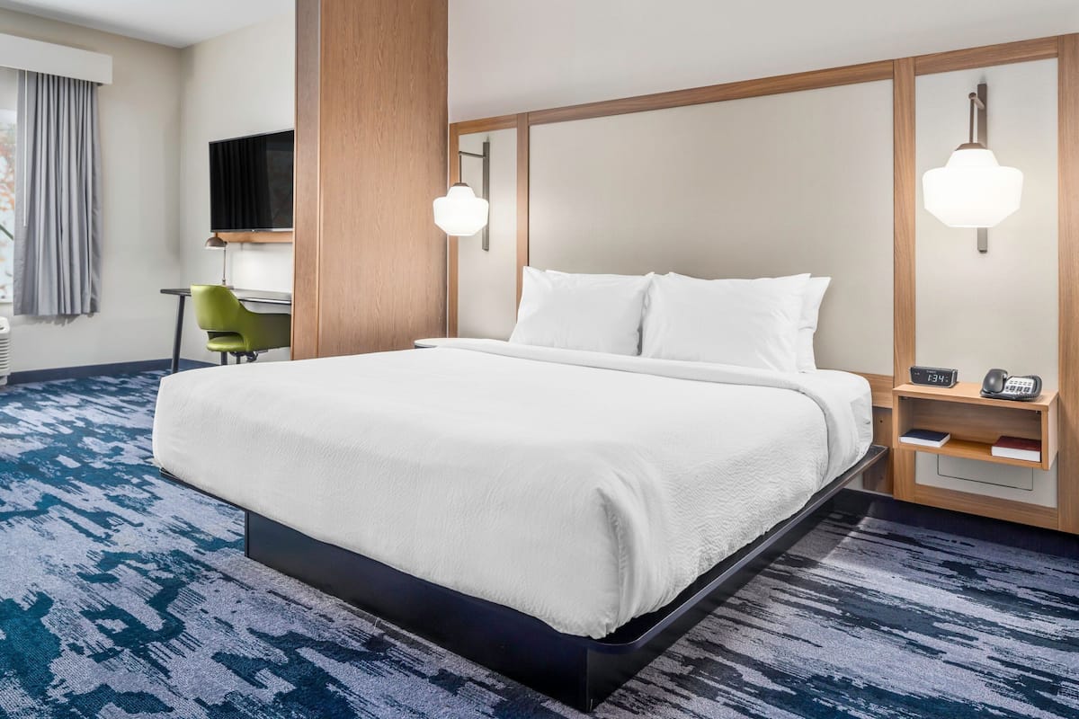 A hotel room with a large bed, a desk, a chair, a TV, and a bedside table with a lamp.