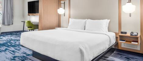 A hotel room with a large bed, a desk, a chair, a TV, and a bedside table with a lamp.
