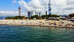 A beach with people swimming and sunbathing, a tall tower, and modern buildings in the background. - Orbi City Batumi Apartment With sea Views (Batumi)