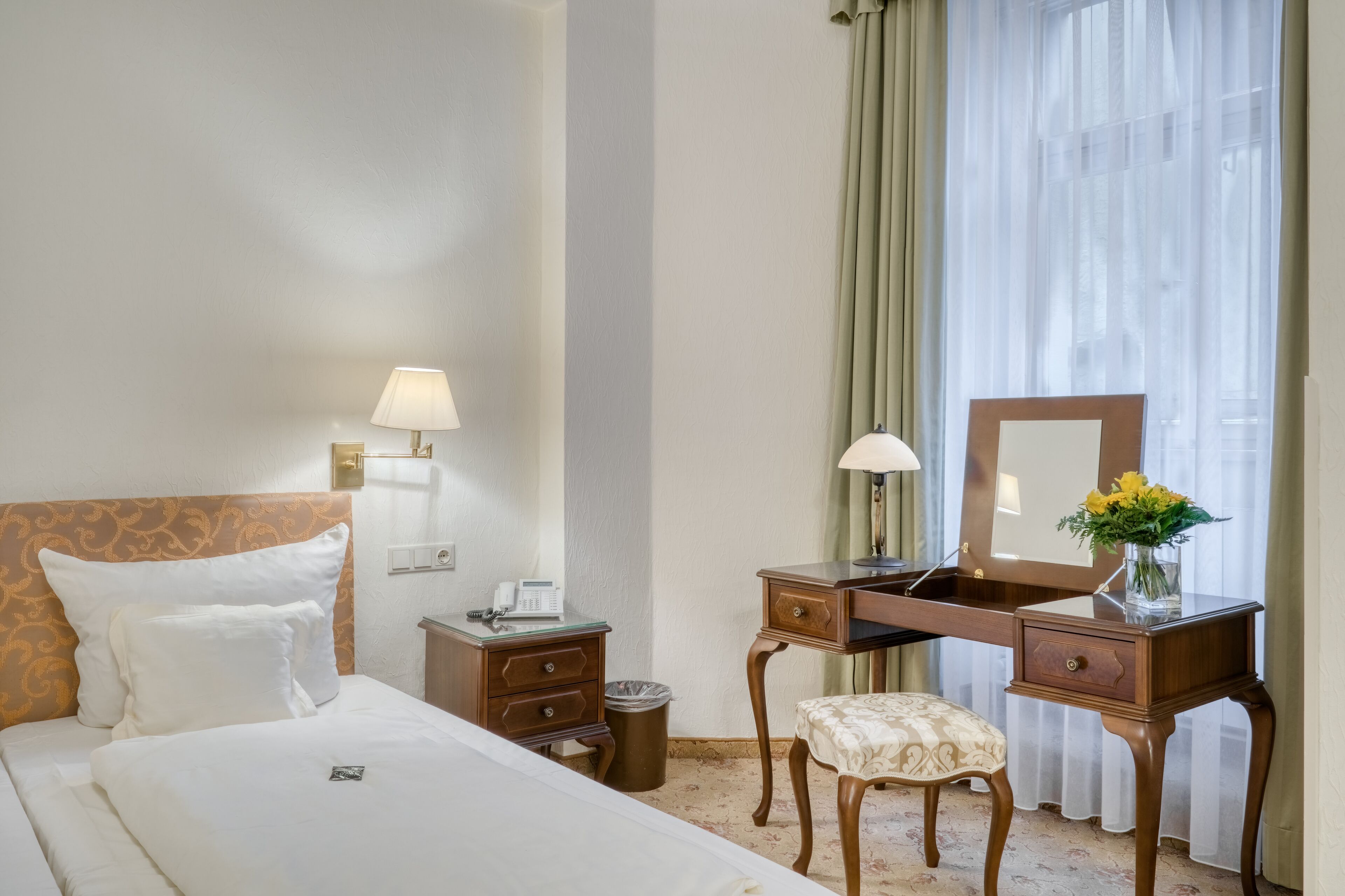 A hotel room with a bed, a desk, a chair, a trash bin, and a window with curtains.