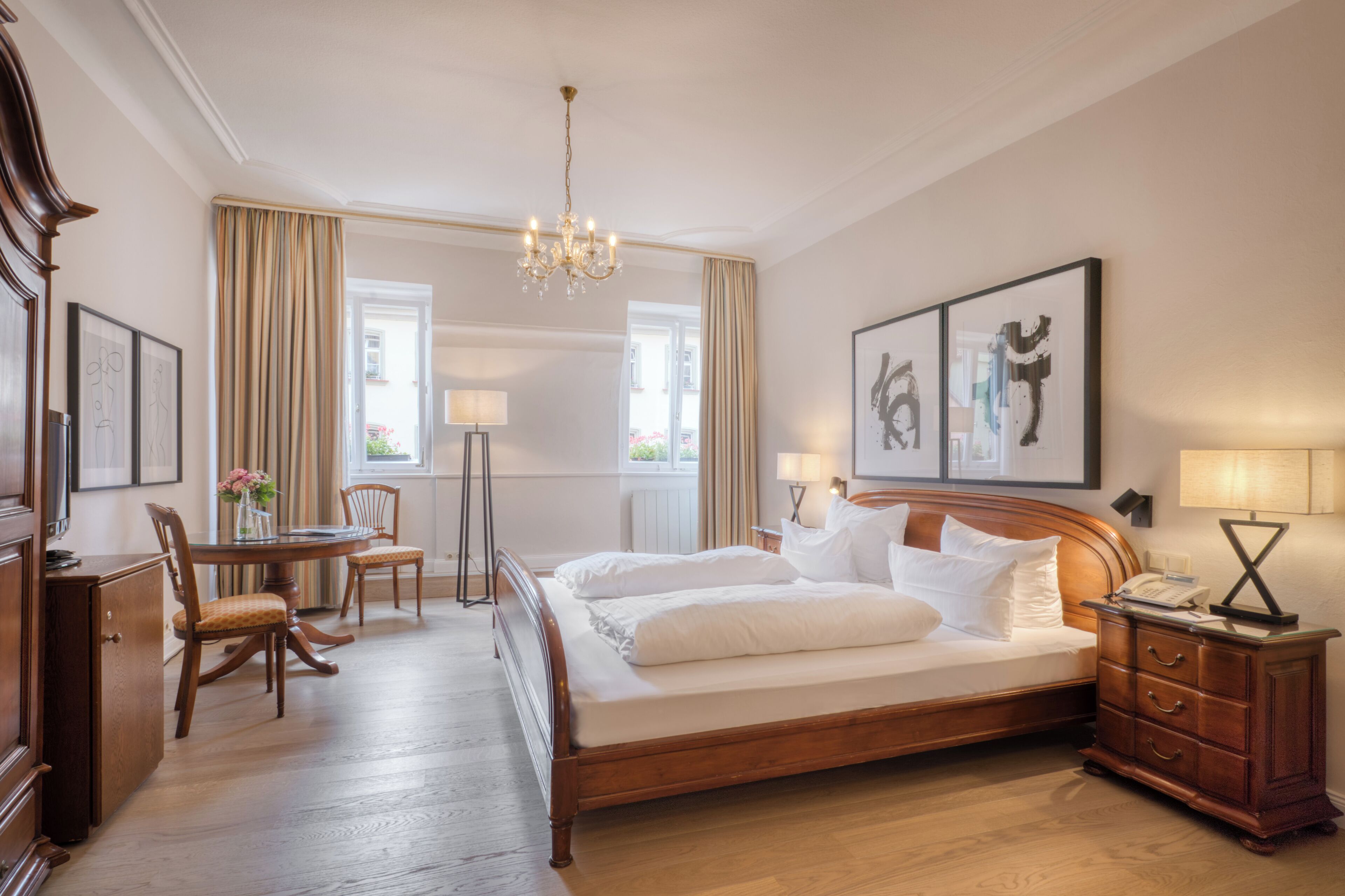 Une chambre d’hôtel avec un grand lit, un bureau avec une chaise, une petite table avec un vase de fleurs et un lustre.