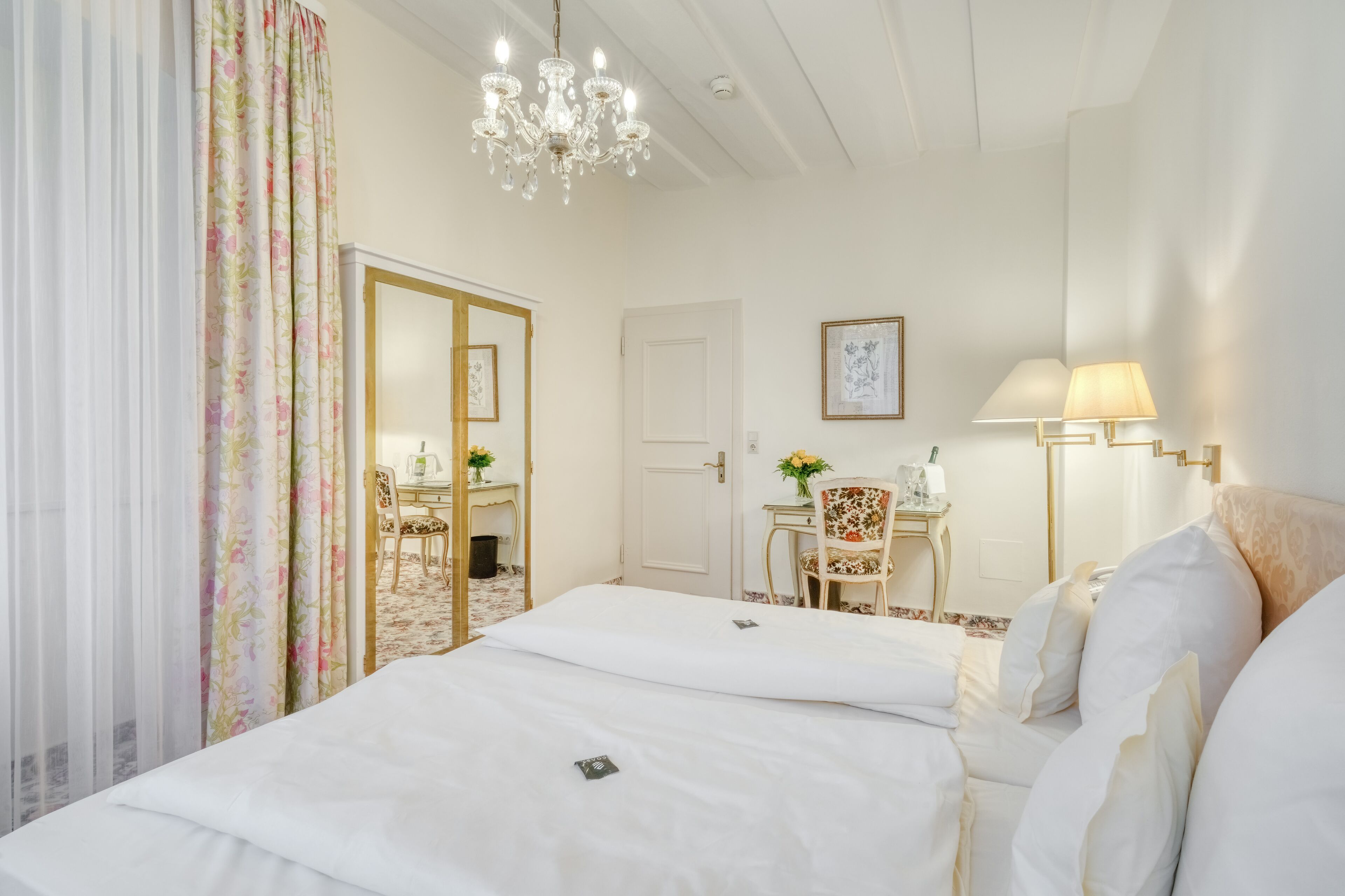 A hotel room with a large bed, a desk, a chair, a lamp, and a chandelier.
