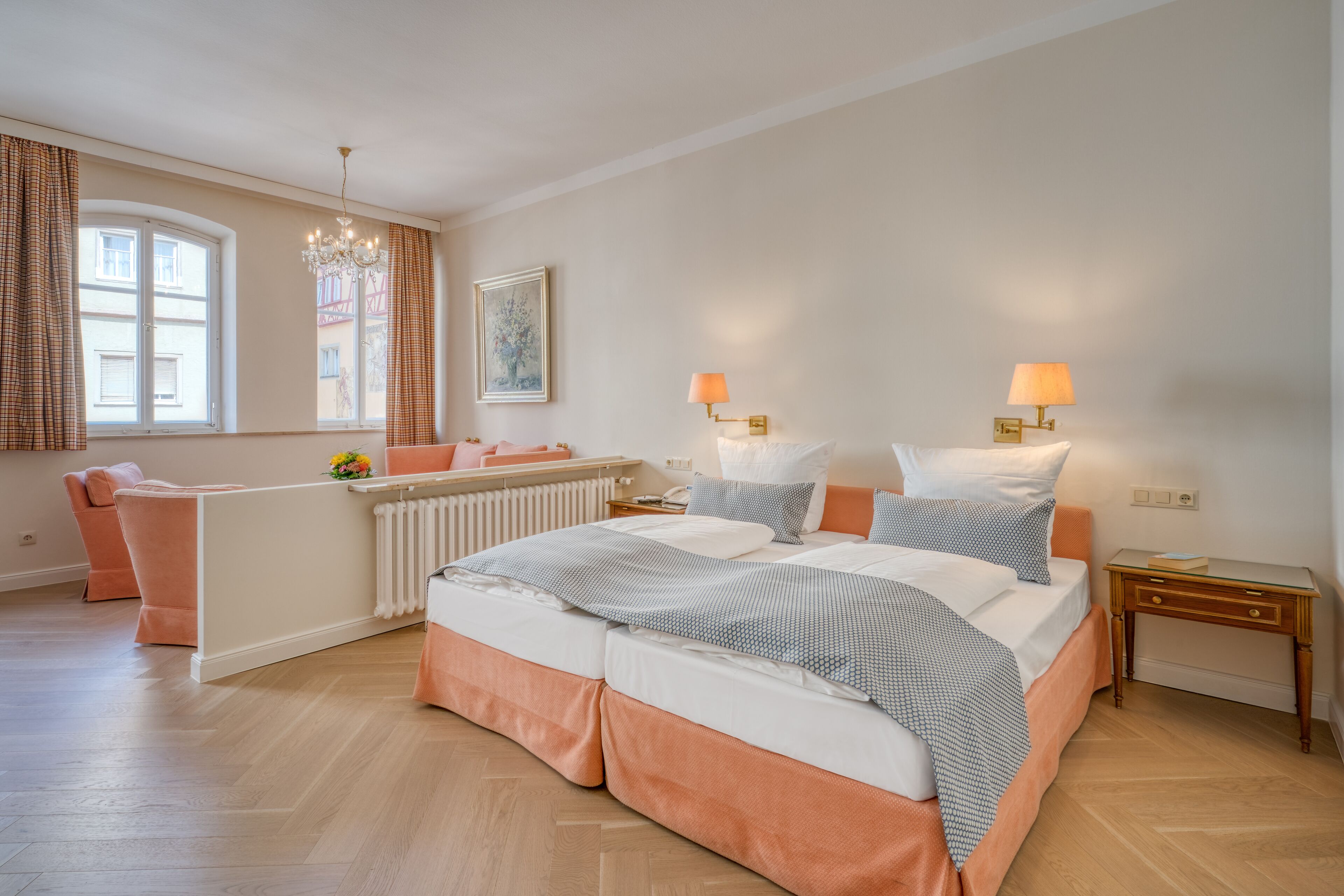 A bedroom with a large bed, two bedside tables with lamps, a radiator, a window with curtains, and a chair.