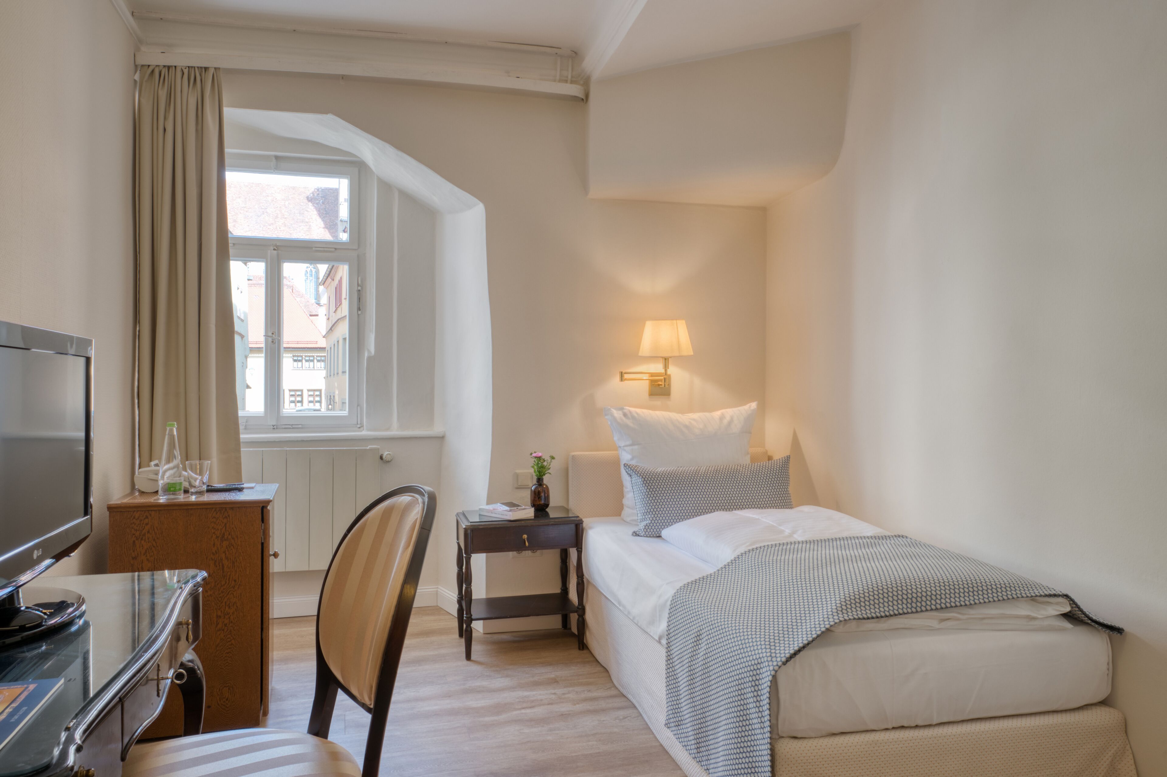 Habitación de hotel con cama, escritorio con televisor, silla, mesita de noche con lámpara y ventana con cortinas.