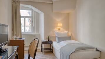 Habitación de hotel con cama, escritorio con televisor, silla, mesita de noche con lámpara y ventana con cortinas.