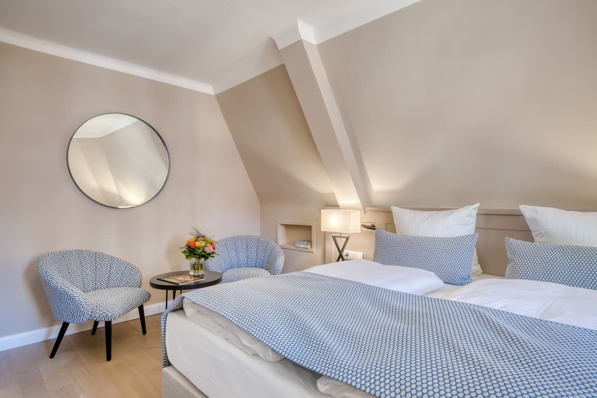 A modern hotel room with a large bed, a round mirror, a small table with a vase of flowers, and two patterned chairs.