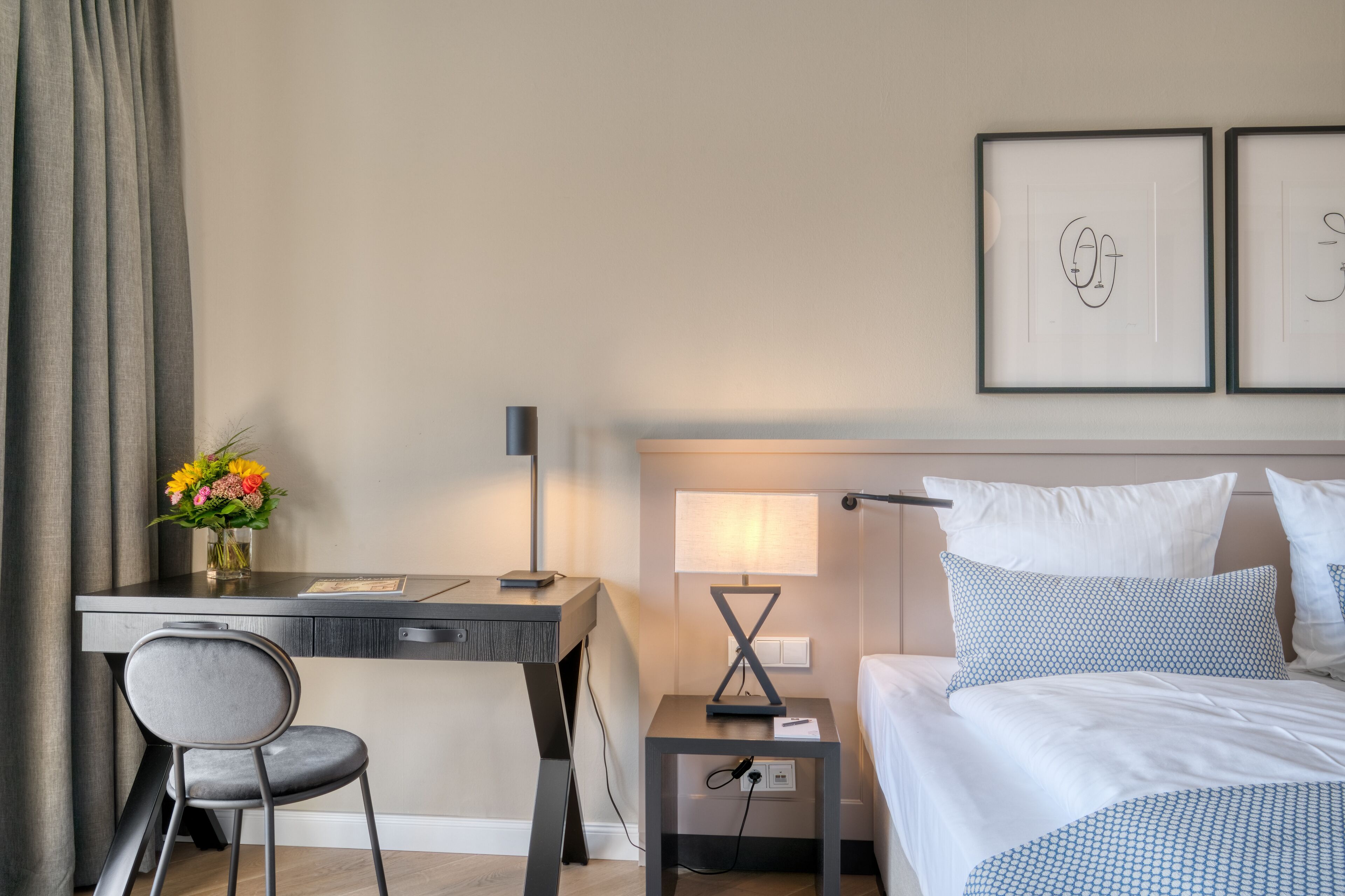 A hotel room with a bed, a desk, a chair, a nightstand, a lamp, and a framed artwork on the wall.