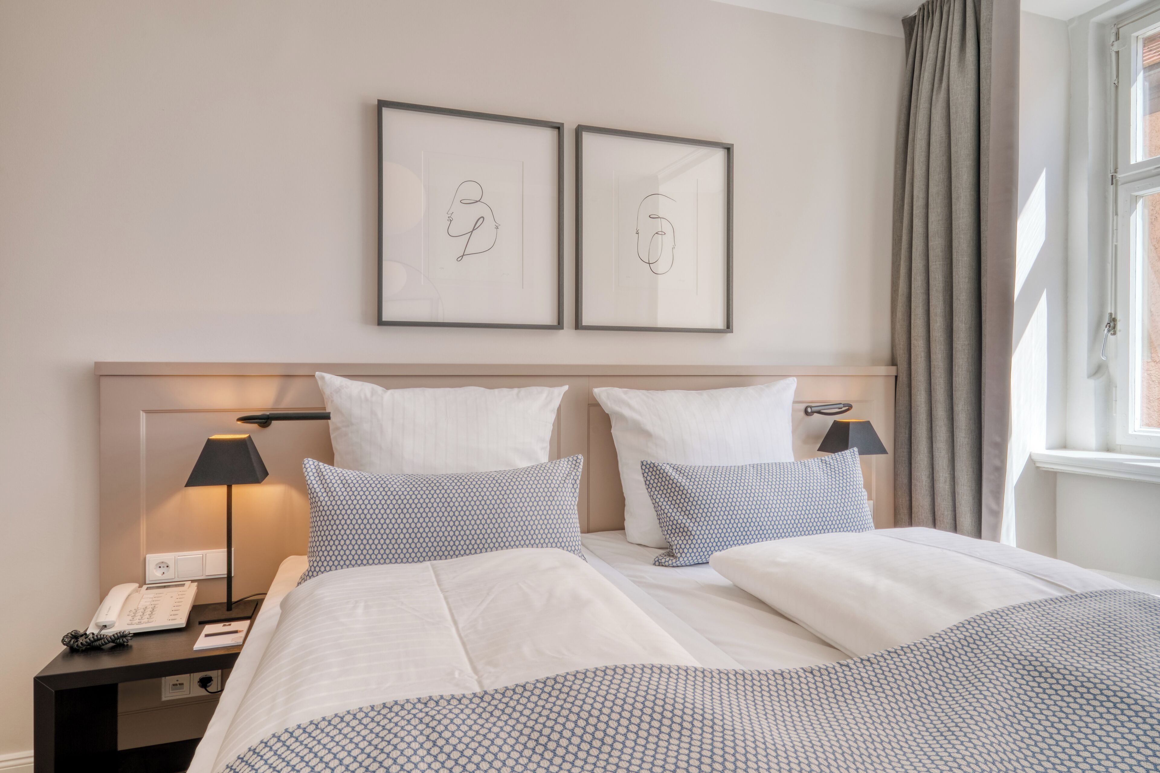 Ein Hotelzimmer mit einem Bett, zwei Nachttischen, einer Lampe, einem Telefon und zwei gerahmten Kunstwerken an der Wand.