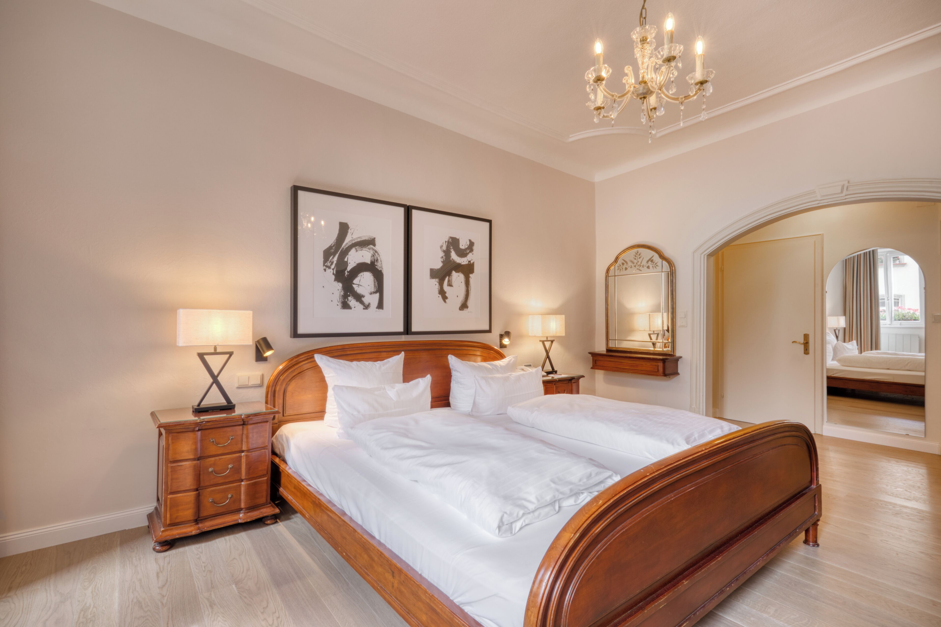 A spacious bedroom with a large bed, wooden headboard, two bedside tables with lamps, and a chandelier.