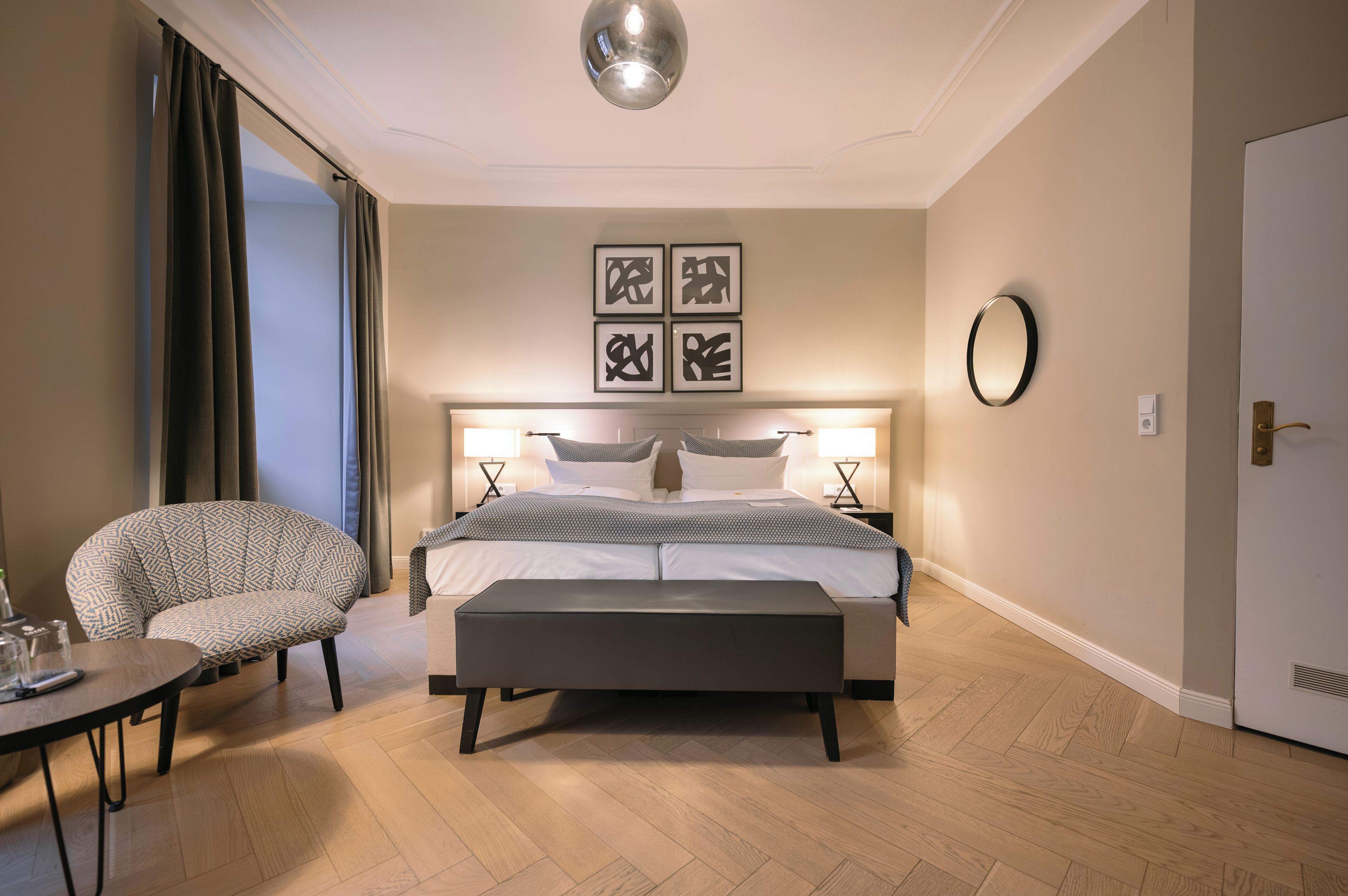 A hotel room with a bed, bedside tables, a chair, a round mirror, and a door.