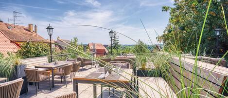 Un patio exterior con mobiliario de mimbre y mesas, rodeado de follaje y vistas a un paisaje urbano.