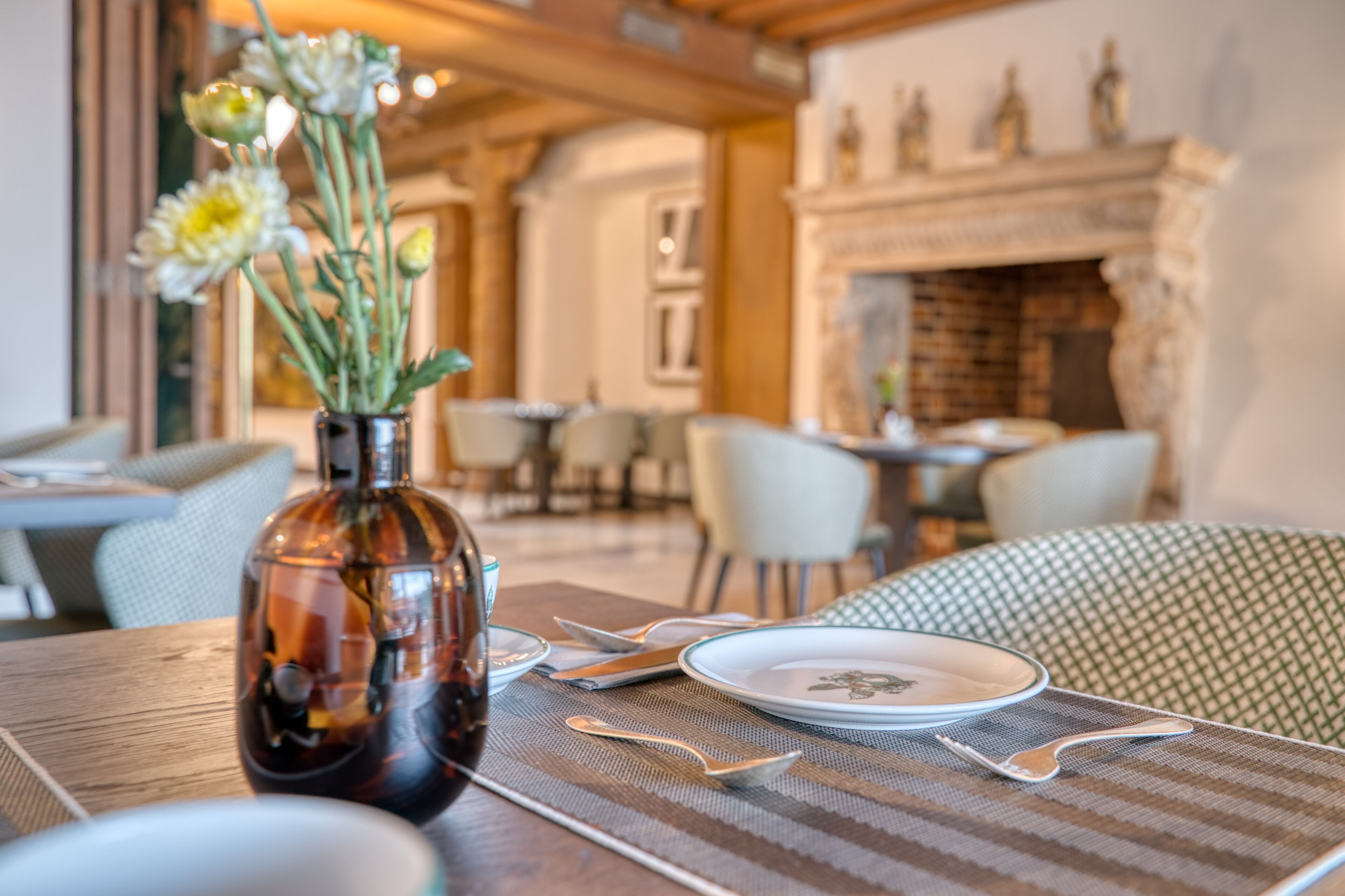 A dining area with a table set for two, a vase with flowers, and a fireplace in the background.