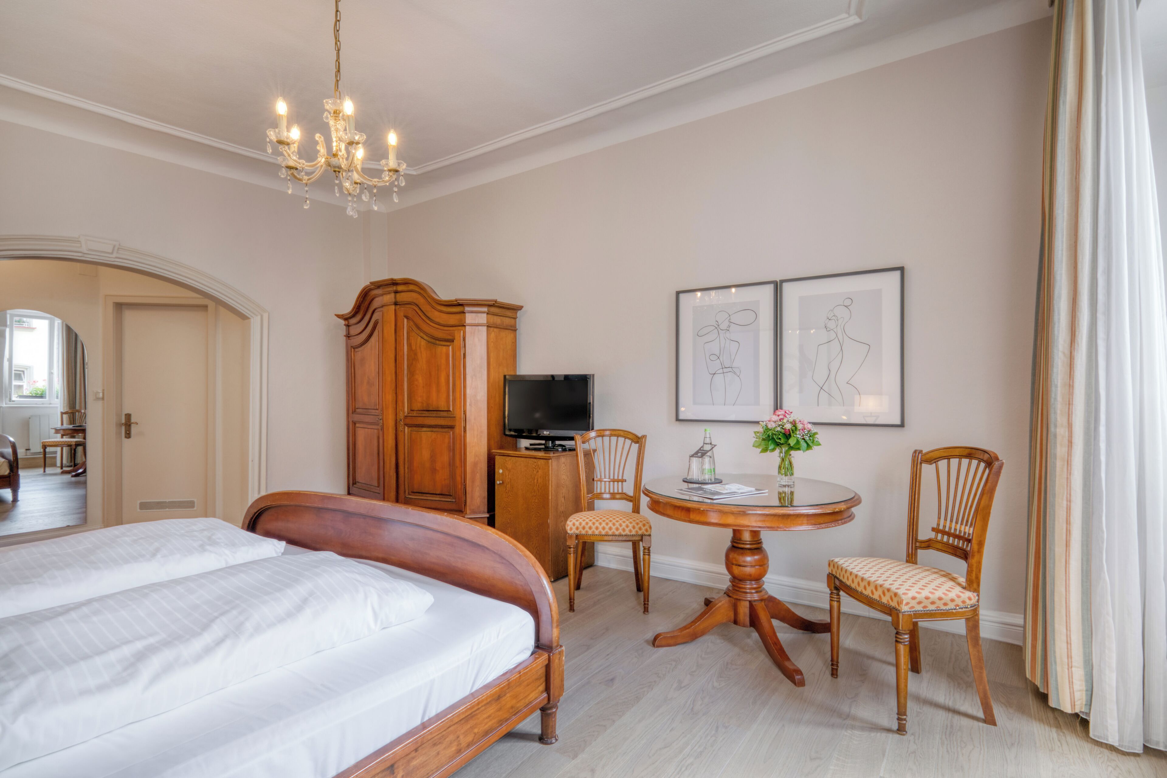 A hotel room with a large bed, a wooden wardrobe, a television, a dining table with chairs, and artwork on the wall.