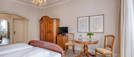 A hotel room with a large bed, a wooden wardrobe, a television, a dining table with chairs, and artwork on the wall.