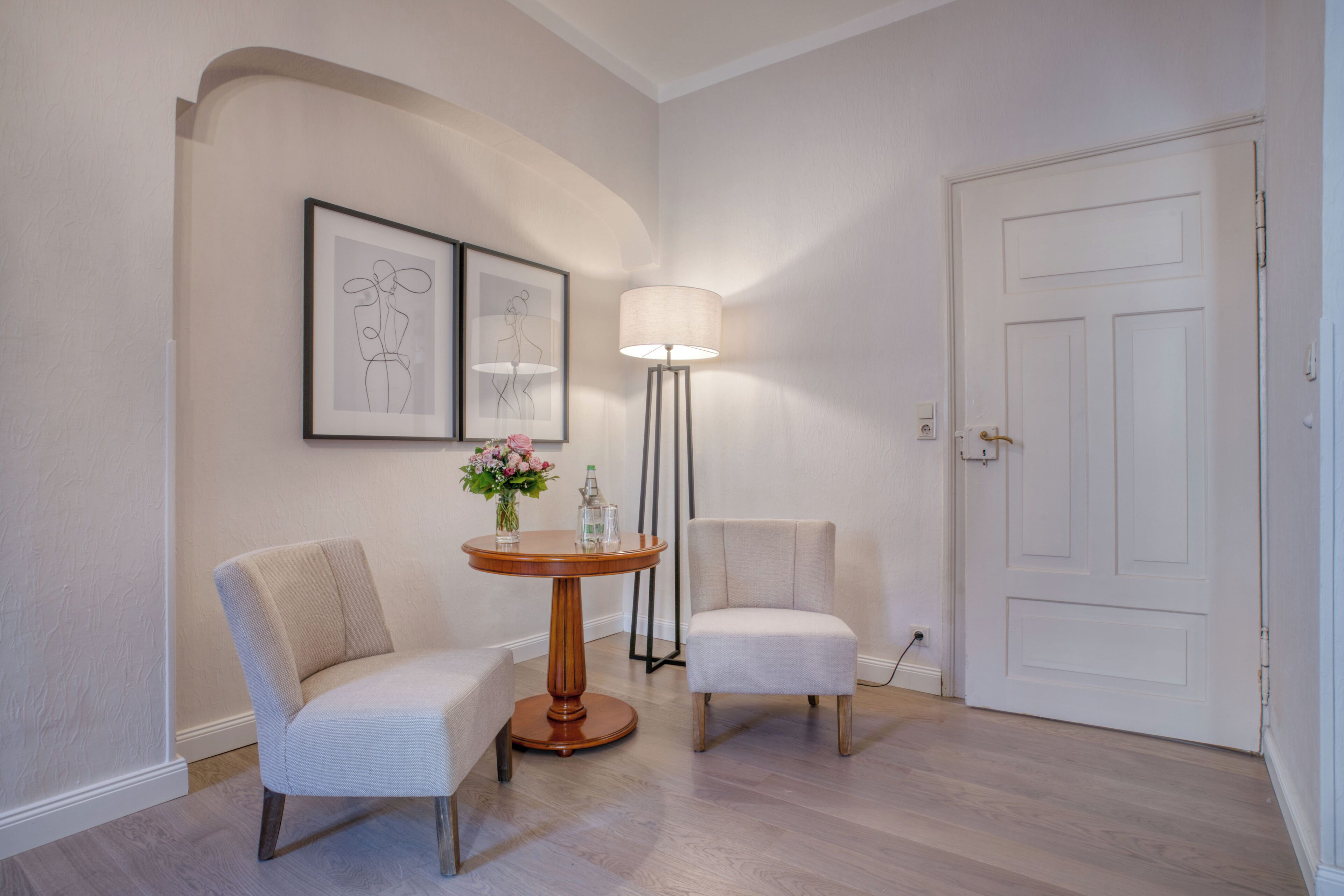 A room with two chairs, a round table with a vase of flowers, a floor lamp, and framed artwork on the wall.
