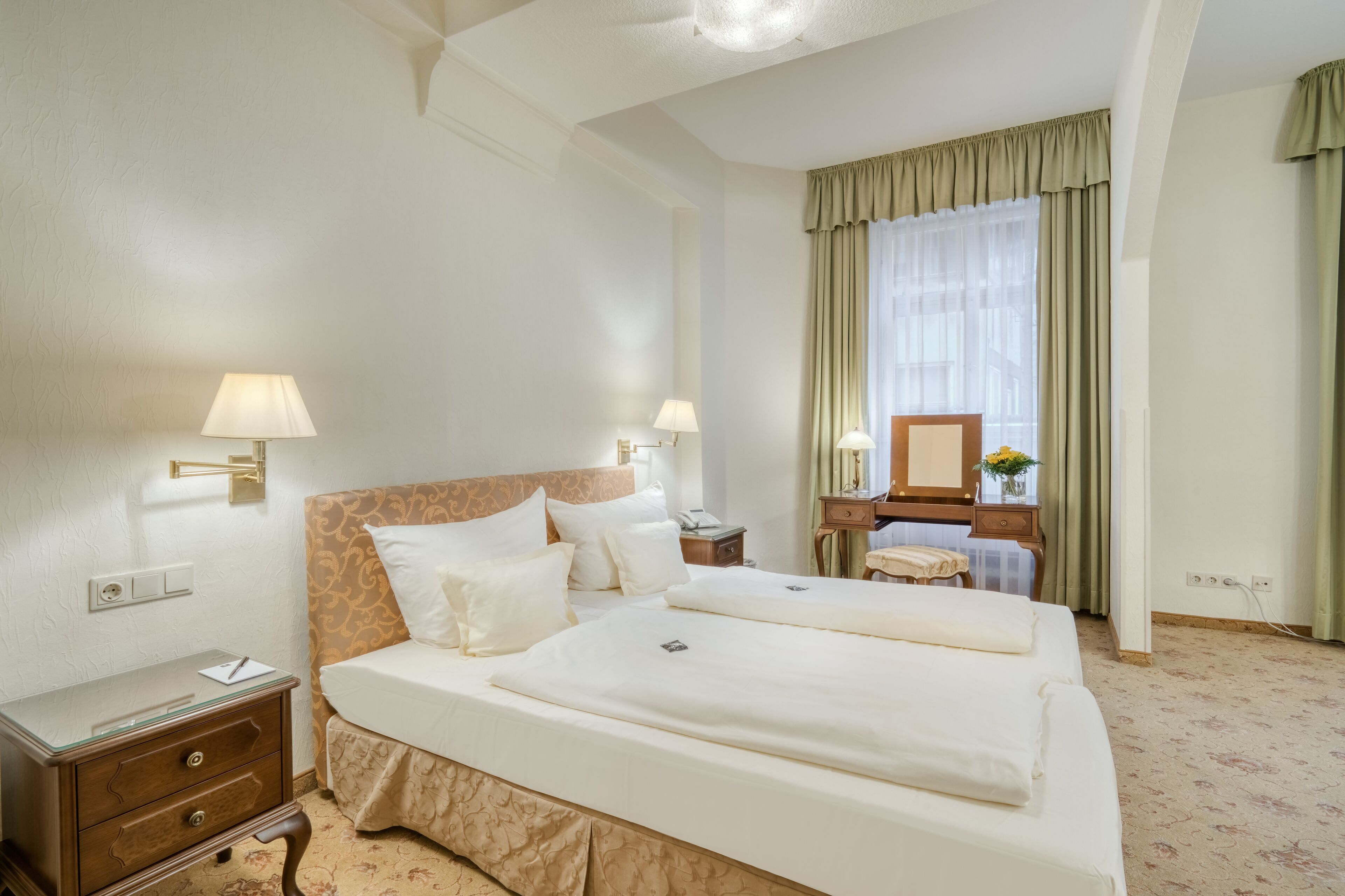 A hotel room with a large bed, two bedside tables, a nightstand, a mirror, and a window with curtains.