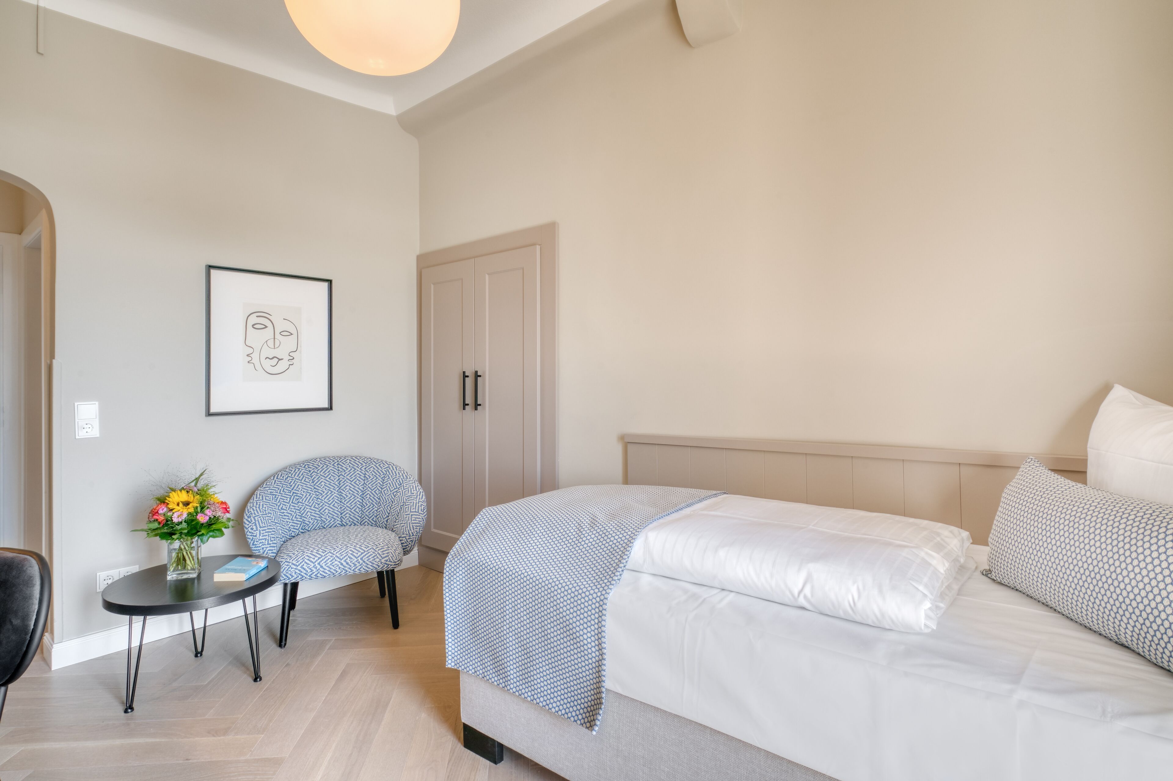 Ein modernes Hotelzimmer mit einem großen Bett, einem kleinen Tisch mit Blumenvase, einem Sessel und einem gerahmten Kunstwerk an der Wand.