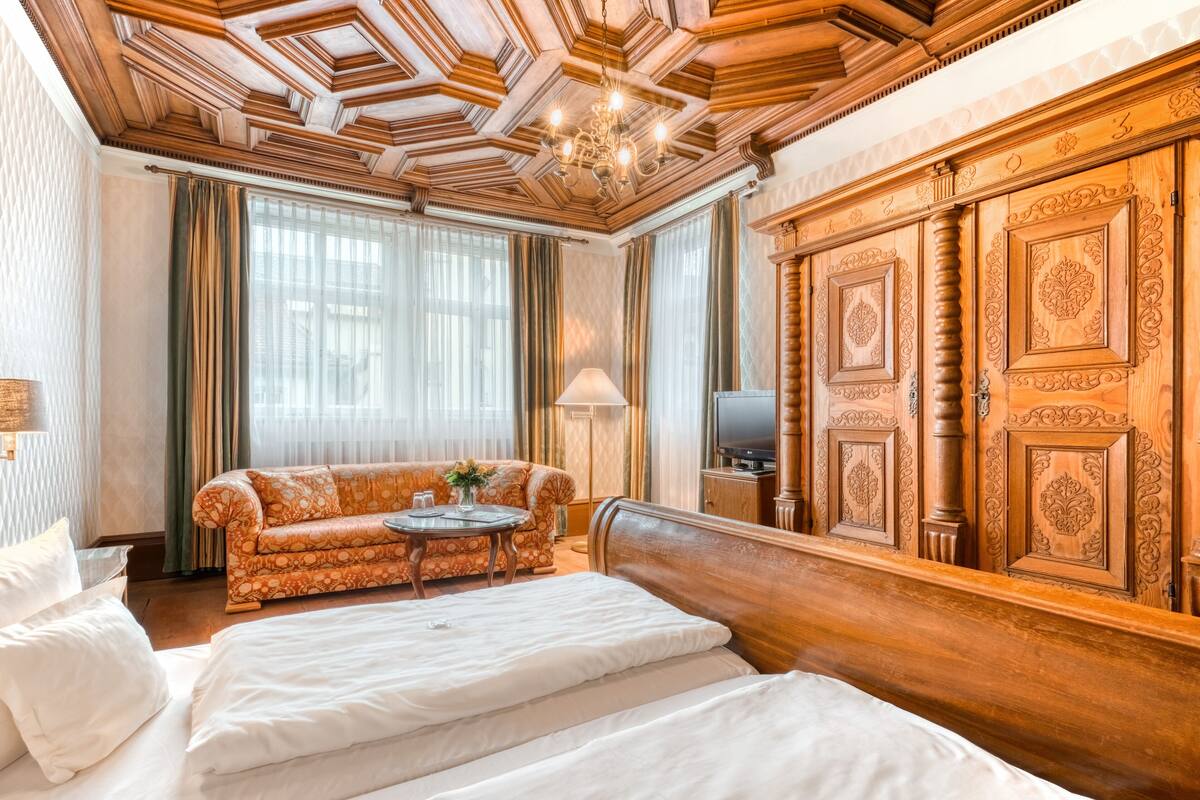 A hotel room with a wooden ceiling, a large bed, a sofa, a TV, and a wooden wardrobe.