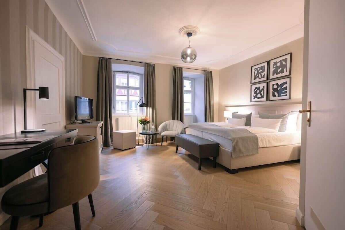 A hotel room with a bed, desk, chair, TV, and artwork on the wall.