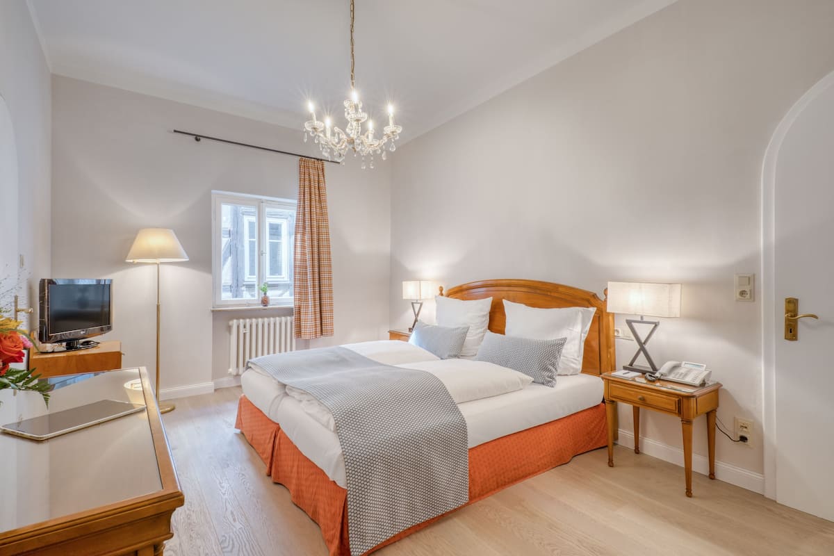 A hotel room with a large bed, bedside tables, a TV, and a chandelier.
