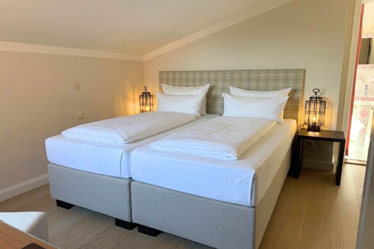A double bed with white linens, a headboard with a plaid pattern, two bedside tables with lamps, and a wooden floor.