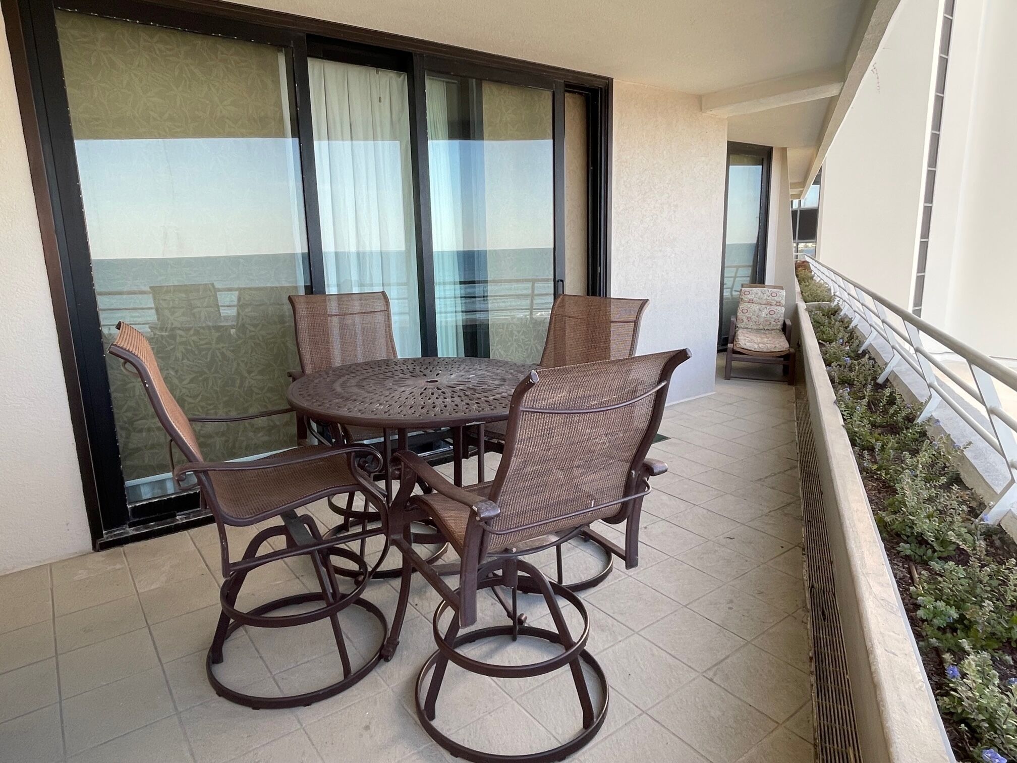 Een balkon met een ronde tafel, vier rieten stoelen, een kleine bank en een railing met planten.