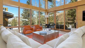 A spacious living area with large windows, a leather sofa, a coffee table, and a view of trees and mountains. - Modern Vail Winter Escape by InvitedHome | Walk to Ski Shuttle, Hot Tub (Vail)