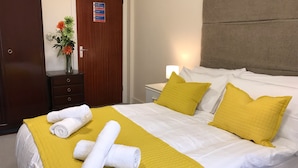 Habitación de hotel con cama, dos almohadas, mesita de noche, lámpara, cómoda, espejo y puerta con un cartel.