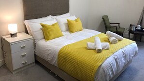 Una cama bien hecha con ropa de cama blanca y amarilla, una lámpara de noche y una silla en la esquina.
