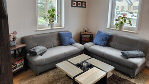 A modern living room with a grey sectional sofa, a wooden coffee table, and two windows with potted plants.