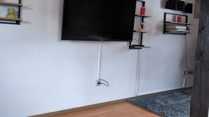A room with a mounted flat-screen TV, a bookshelf with books and decorative items, a small yellow vase, and a blue rug.