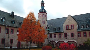 Un bâtiment historique doté d’une tour imposante, de fenêtres à cadre rouge et d’une cour intérieure où le feuillage d’automne crée un tableau magnifique.