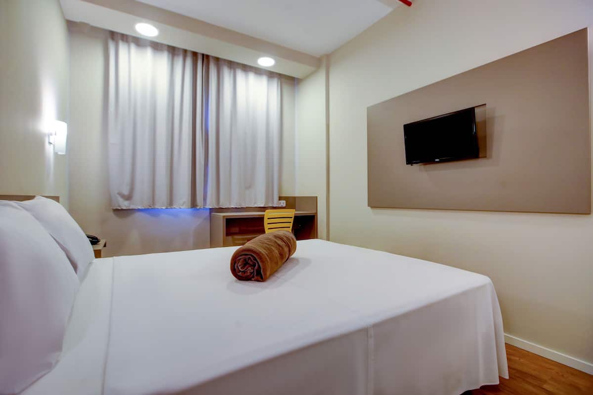 A hotel room with a bed, a TV mounted on the wall, a desk with a chair, and a window with curtains.