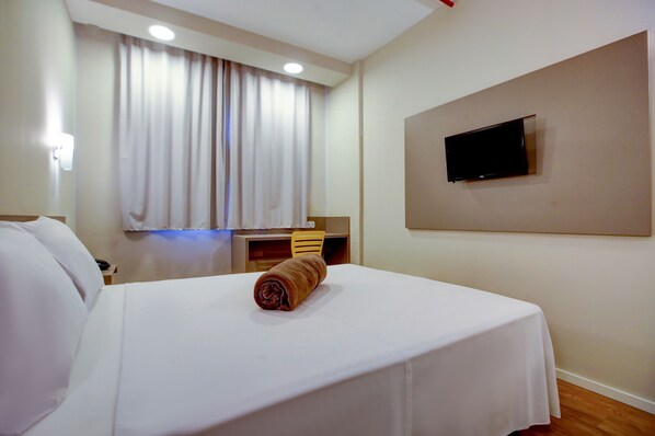 A hotel room with a bed, a TV mounted on the wall, a desk with a chair, and a window with curtains.
