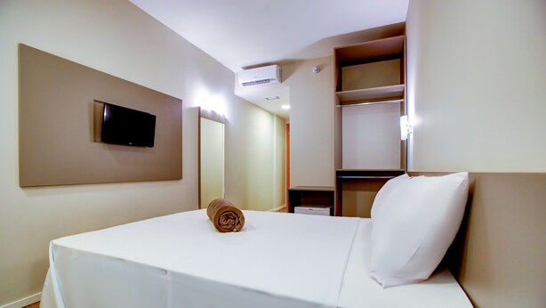 Une chambre d’hôtel avec un lit, un téléviseur fixé au mur, un miroir et une étagère sur laquelle sont posés quelques objets.