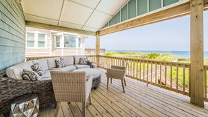 Terraza techada con un sofá y sillas de mimbre, con vista a una playa y el océano.