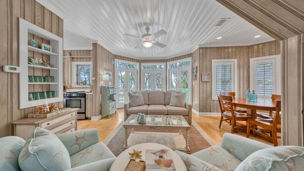 A spacious living room with a large sofa, a coffee table, and a dining area with a wooden table and chairs. There is a kitchen area with a stove and cabinets, and large windows providing natural light. - Seaside "Frose Rose" | 2 BR | Sleeps 6 | Private | Steps to Beach! (Santa Rosa Beach)