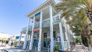 A two-story building with a balcony, a sign reading "SAND DOG BOOKS", and a palm tree to the right. - Seaside "Frose Rose" | 2 BR | Sleeps 6 | Private | Steps to Beach! (Santa Rosa Beach)