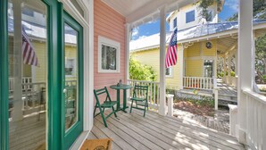 A porch with green chairs and a small table, a door with a glass panel, and two houses with American flags. - Seaside "Frose Rose" | 2 BR | Sleeps 6 | Private | Steps to Beach! (Santa Rosa Beach)