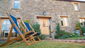 Un bâtiment en pierre avec une porte en bois, deux fenêtres et un petit jardin agrémenté de plantes en pot et d’une pelouse.