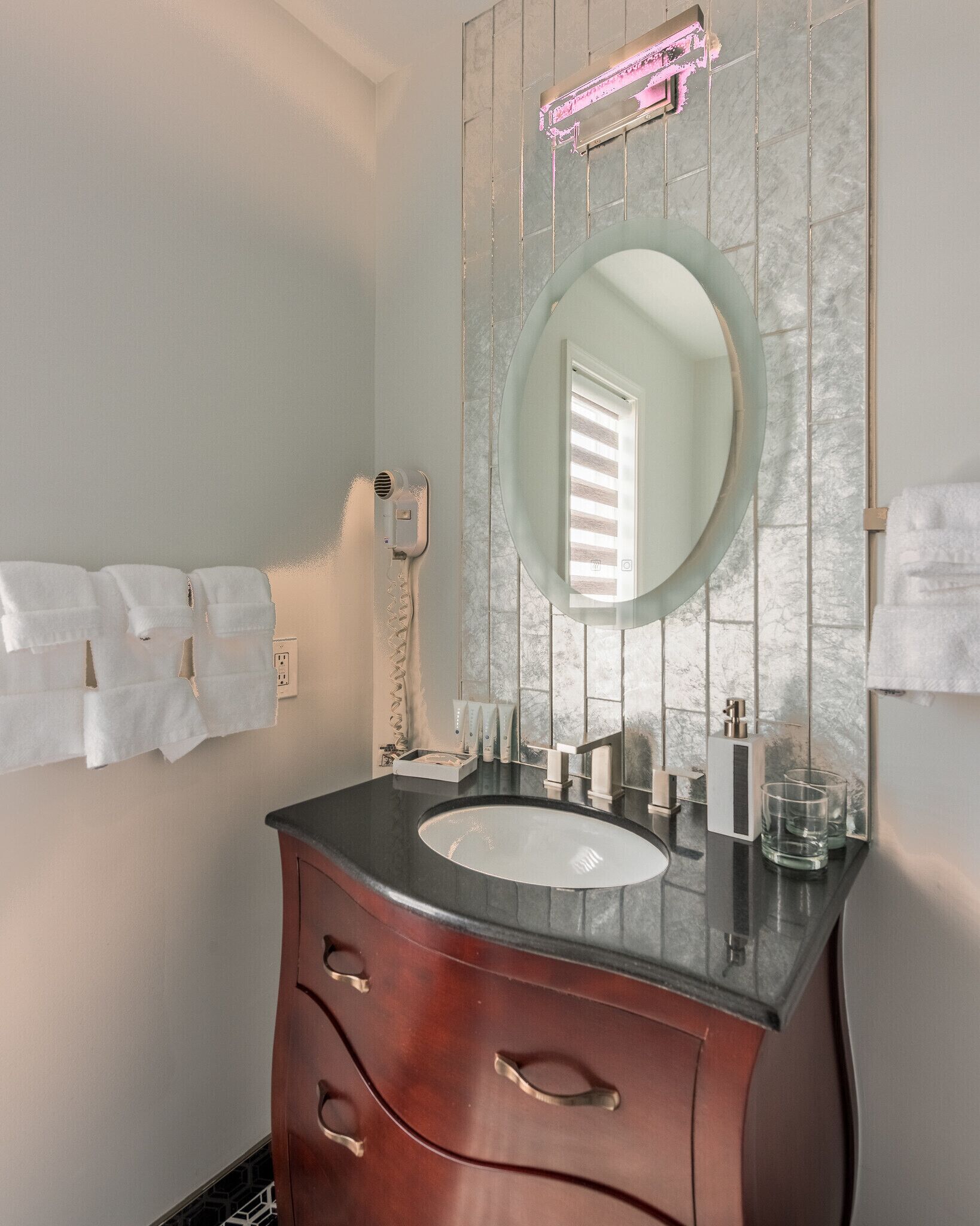 A bathroom with a marble backsplash, a round mirror, a sink, a vanity with drawers, and a towel rack.