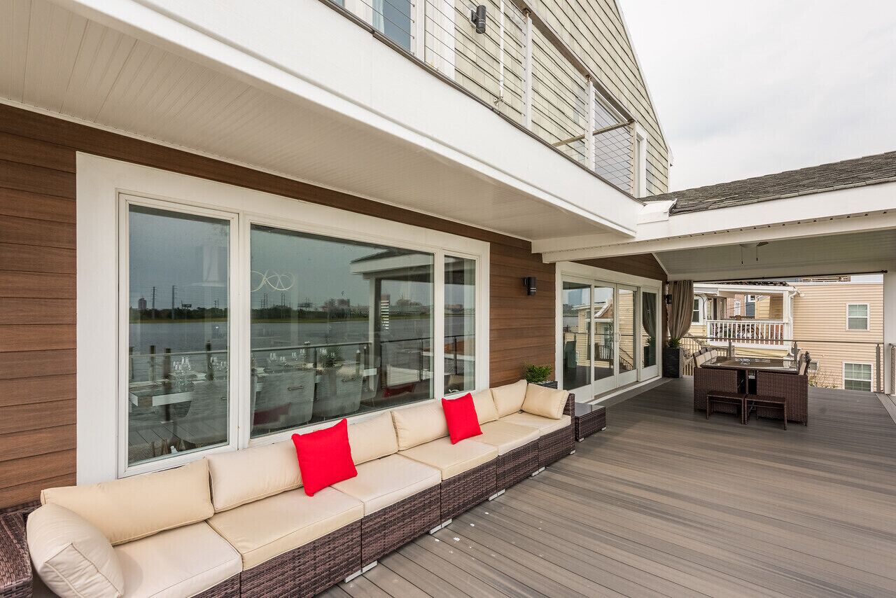 A covered outdoor area with a long wicker sofa and a table, large windows, and a view of the water.