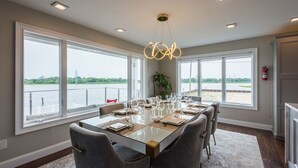 Un comedor con una mesa grande dispuesta para seis personas, ventanas amplias con vista a una masa de agua y una lámpara de araña moderna.