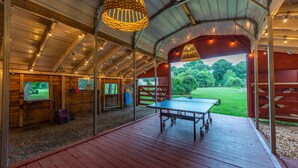 Un espacio al aire libre techado con mesa de ping pong, bancos de madera y vista a un campo verde.