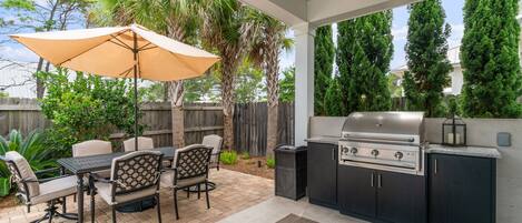 An outdoor kitchen with a grill, dining table, chairs, and an umbrella.
