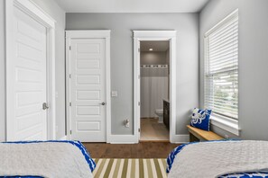A bedroom with two beds, a window with blinds, a door leading to a bathroom, and a small bench with a pillow. - Golf Cart ~ Private Pool ~ Near Beach ~ Grill (Santa Rosa Beach)